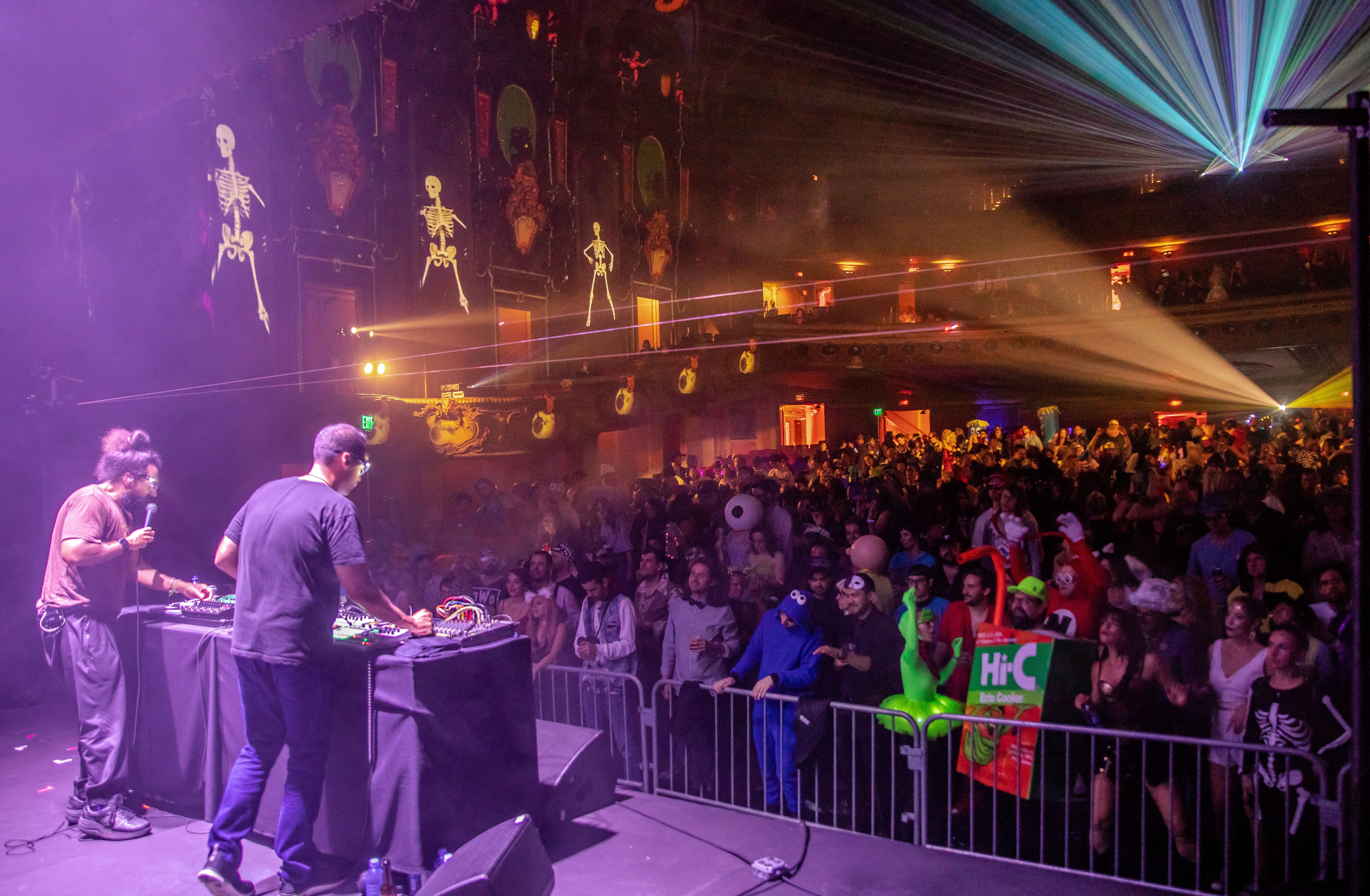 two DJs on a stage in a theatre in front of a crowd of people in costumes such as a Hi-C, Cookie Monster, a minion, and Kurt Cobain an d a skeleton