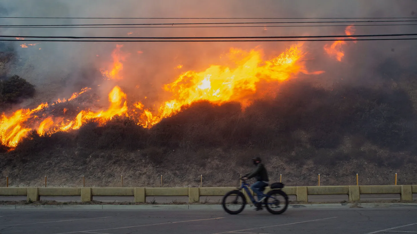 A person rides a bike as the Palisades Fire burns during a windstorm on the west side of Los Angeles, California, U.S. January 7, 2025.