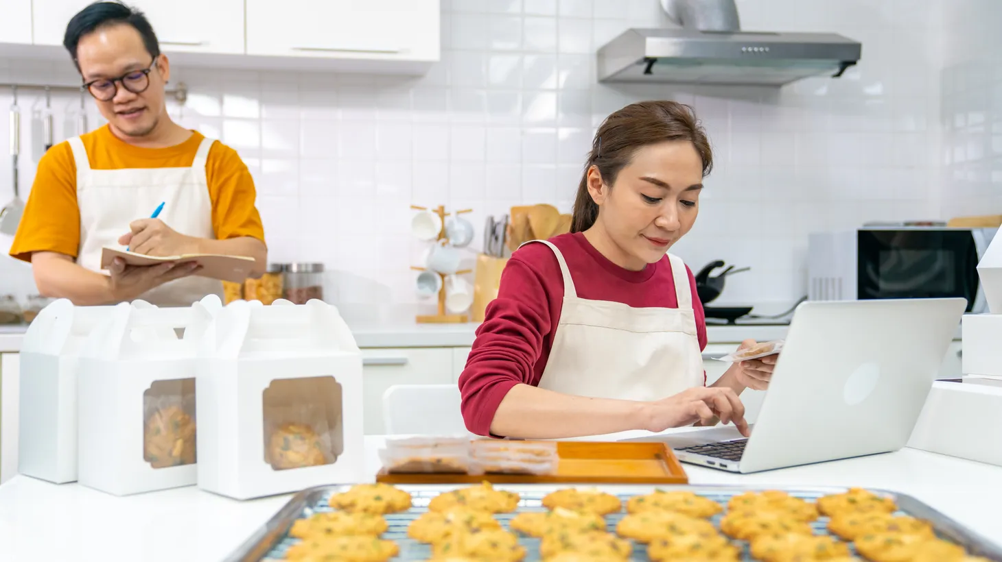 A woman processes customers’ orders on a laptop as a man takes notes for their home bakery business.